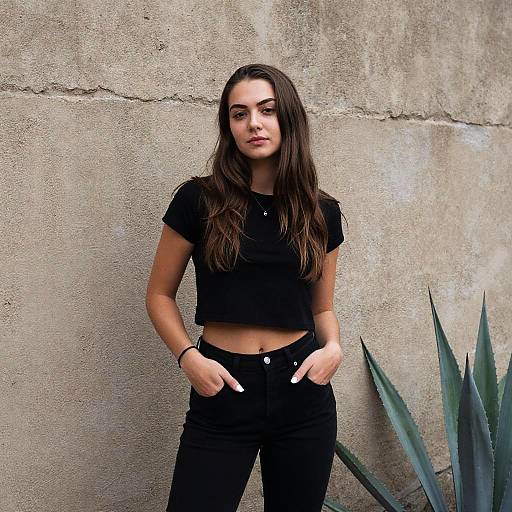 Photograph of a young woman with long brown hair, wearing a black crop top and jeans, hands in pockets, standing against a textured concrete wall with