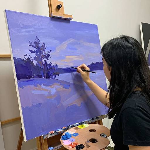Asian woman with black hair painting a blue-toned landscape on canvas, using a brush, in an artist's studio with palette.
