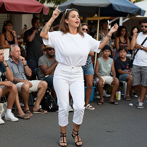 Photograph of a brunette woman in white outfit and black sandals, standing and gesturing with arms raised, in front of a seated audience under colorful umb