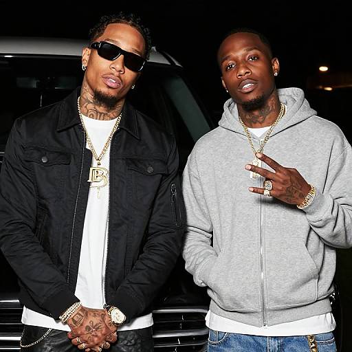 Photograph of two African-American men wearing sunglasses, gold chains, and casual clothing, standing in front of a dark car.