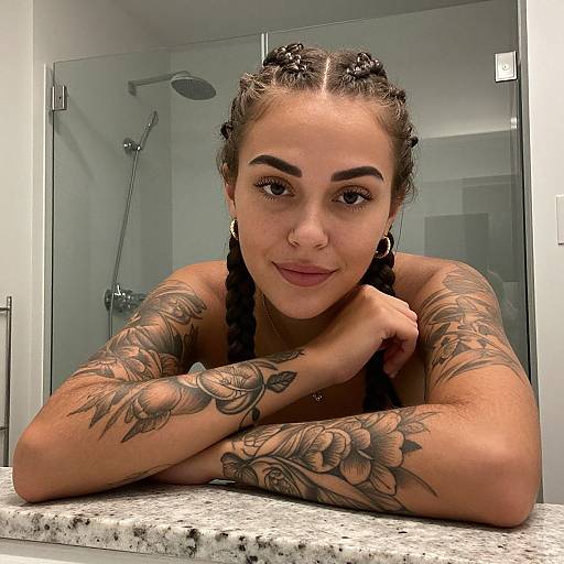 Tattooed woman with braided hair and hoop earrings leans on bathroom counter, smiling; shower in background, modern bathroom setting. Photographic image