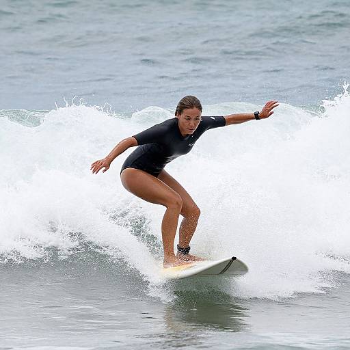 Photograph of a woman in a black wetsuit surfing a wave, her right arm extended, left arm bent, splashing water beneath her white