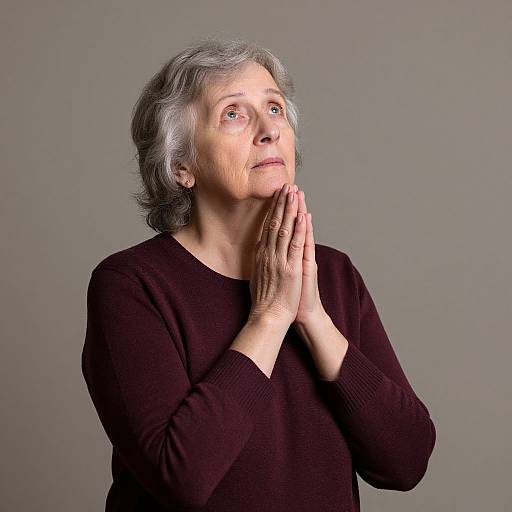 Photograph of an elderly woman with gray hair, wearing a black sweater, hands clasped in prayer, looking upward with a thoughtful expression against a plain