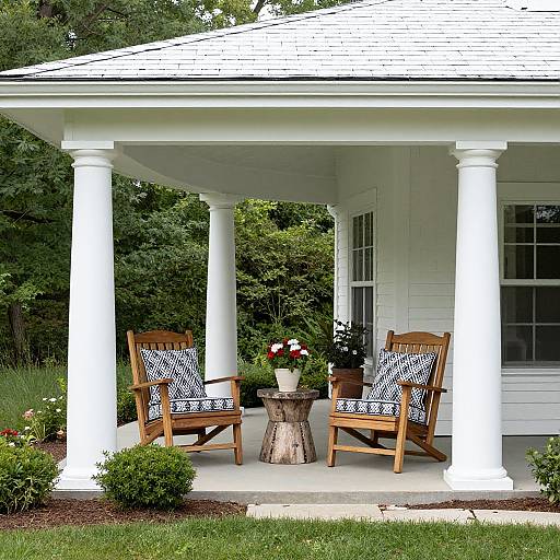 Photograph of a white-painted porch with two wooden chairs, black-and-white patterned cushions, small wooden table, and potted flowers. Background