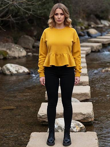 Photograph of a woman with wavy brown hair, wearing a yellow long-sleeve top, black pants, and black shoes, standing on a