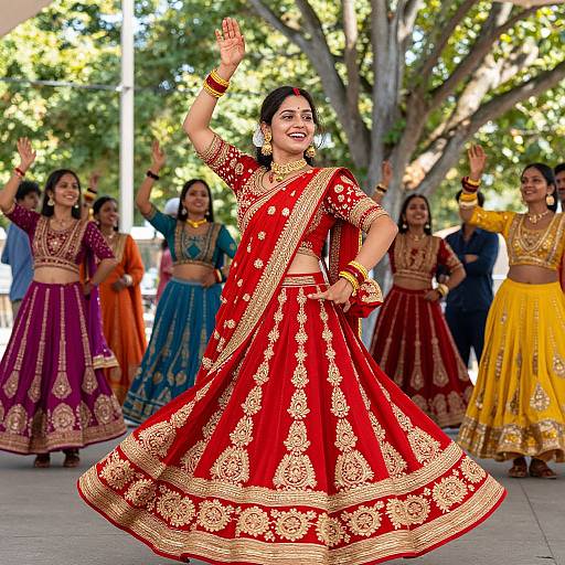 Photograph of a smiling Indian woman in a vibrant red and gold traditional lehenga, dancing with friends in colorful attire, outdoors under trees.