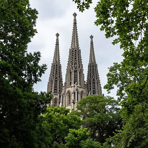 Photograph of a Gothic-style cathedral with three tall, intricately detailed spires, partially obscured by lush green trees against a bright, cloudy sky.