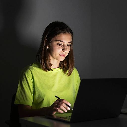Photograph of a young woman with straight brown hair, wearing a bright yellow shirt, typing on a laptop in dim lighting.
