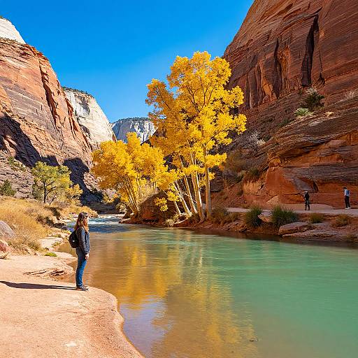 Photograph of a vibrant canyon scene with bright yellow trees, clear turquoise water, and red rock cliffs under a vivid blue sky. A person stands on
