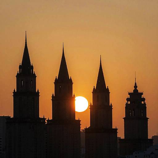 Photograph of a sunset with the sun partially obscured by four silhouetted Gothic-style towers against an orange sky.