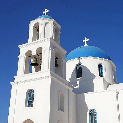 Photograph of a white Greek Orthodox church with blue domes, cross-topped towers, and arched windows against a vibrant blue sky.