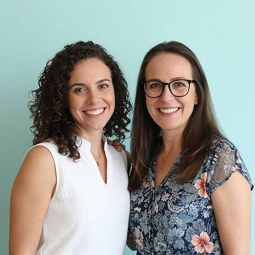 Photograph of two smiling women with curly and straight dark brown hair, wearing white and floral dresses, standing against a light blue background.