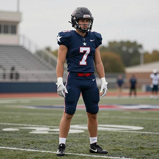 Photograph of a male American football player in navy uniform with white star, number 7, standing on green field, wearing helmet and gloves. Bl