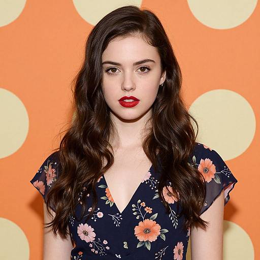 Photograph of a young woman with long, wavy dark brown hair, red lipstick, wearing a black floral dress, against an orange background with large