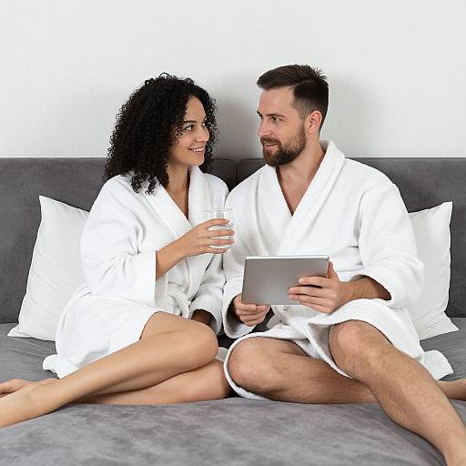 Photograph of a curly-haired woman and bearded man in white bathrobes, sitting on a gray couch, holding drinks and a tablet, smiling