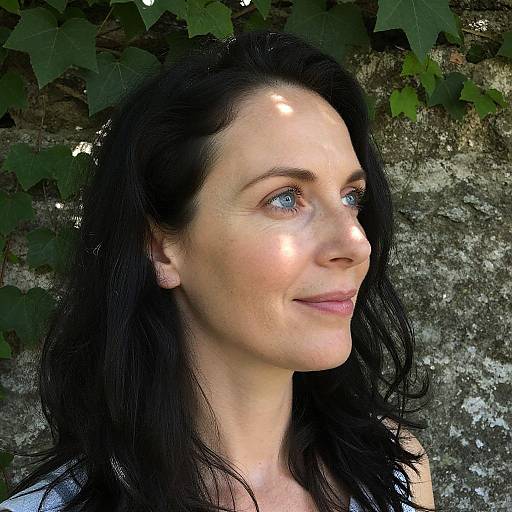 Photograph of a fair-skinned woman with black hair, blue eyes, and subtle smile, standing against a textured stone wall with green ivy leaves