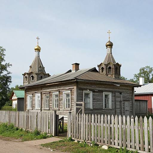 Photograph of a rustic, wooden church with two golden-domed steeples, white picket fence, and overgrown yard on a sunny day.