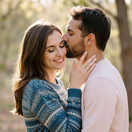 Photograph of a smiling couple kissing softly; woman in blue patterned sweater, man in white shirt, sunlight in background, forest setting.