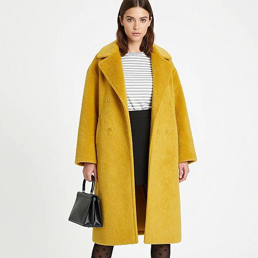 Photograph of a woman with straight brown hair wearing a long yellow wool coat, white-striped shirt, black pants, and black handbag. White background