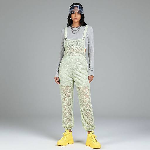 Photograph of a young woman in white lace overalls, grey long-sleeve shirt, yellow sneakers, and black cap, standing against a plain