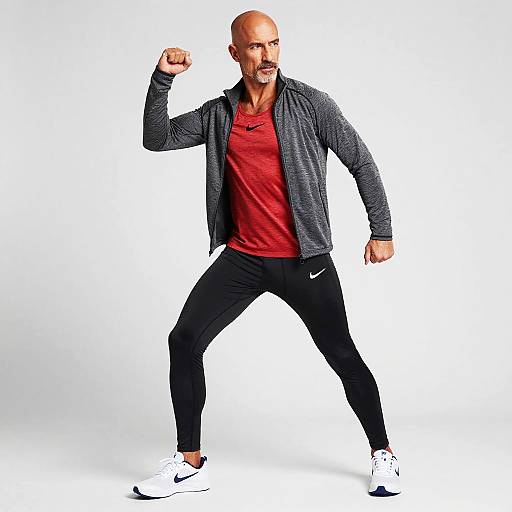 Photograph of a bald, muscular man with a mustache, flexing his right arm, wearing a red shirt, gray cardigan, black Nike
