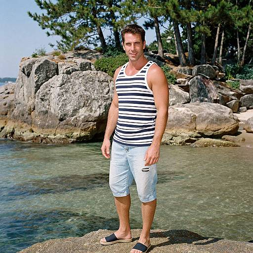 Photograph of a muscular man with short brown hair, wearing a black-and-white striped tank top, white cargo shorts, and black sandals, standing on