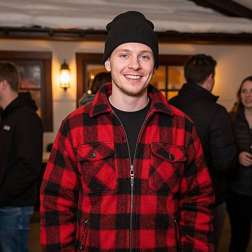 Photograph of a smiling, bearded man in a black beanie and red-black plaid jacket, standing indoors with blurred background. Warm lighting,