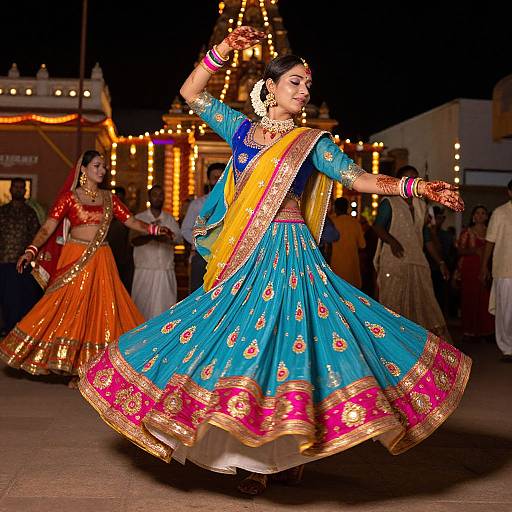 Photograph of two Indian women in vibrant traditional sarees, dancing at night in front of a brightly lit temple, with ornate jewelry and intricate embroidery