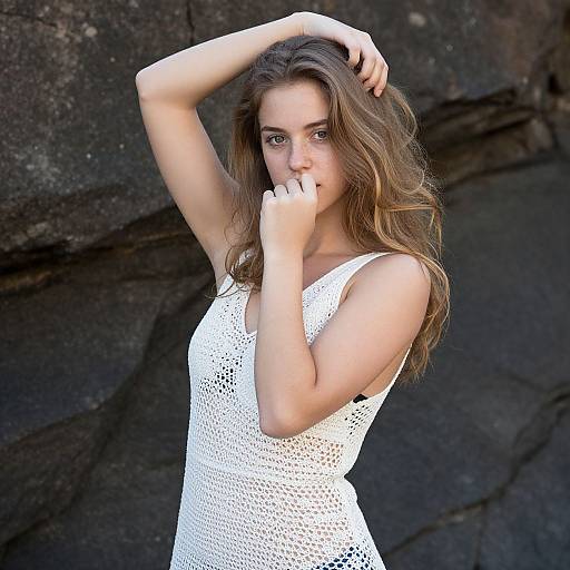 Photograph of a young woman with long brown hair, wearing a white, sleeveless, lace dress, standing against dark rocky background, arms raised,