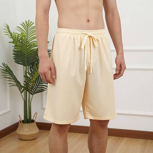 Photograph of a shirtless man wearing light yellow, loose-fitting shorts with a drawstring, standing in a room with a potted plant and