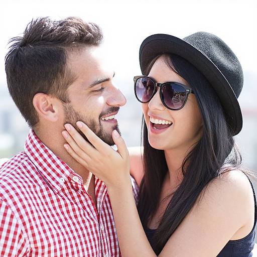 Photograph of a smiling couple; man with dark hair and beard in red checkered shirt, woman with long black hair, black hat, and sunglasses