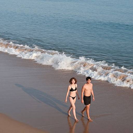 Photograph of a beach scene showing a curly-haired woman in a black bikini and a shirtless man in black swim trunks walking along the wet shoreline