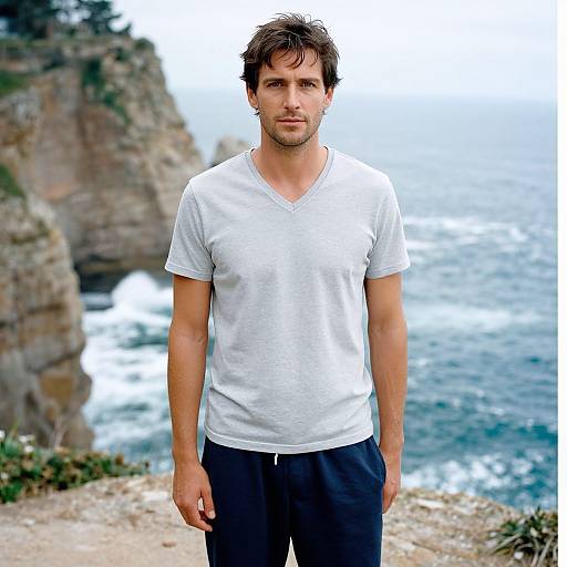 Photograph of a handsome, muscular man with short brown hair, wearing a white V-neck T-shirt and black pants, standing on a rocky coastal cliff