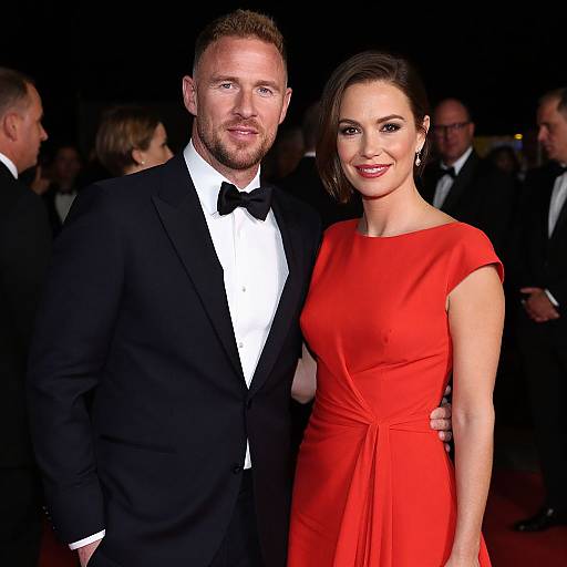 Photograph of a red-carpet event: a bearded man in a black tuxedo and bow tie, standing beside a woman in a vibrant