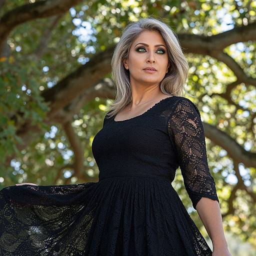 Photograph of a silver-haired woman with green eyes, wearing a black lace dress, standing outdoors against a sunlit tree background.
