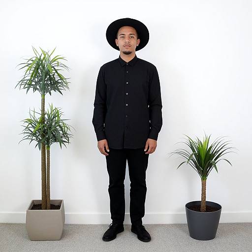 Photograph of a young Asian man in black outfit and hat, standing between two potted plants against a white wall.