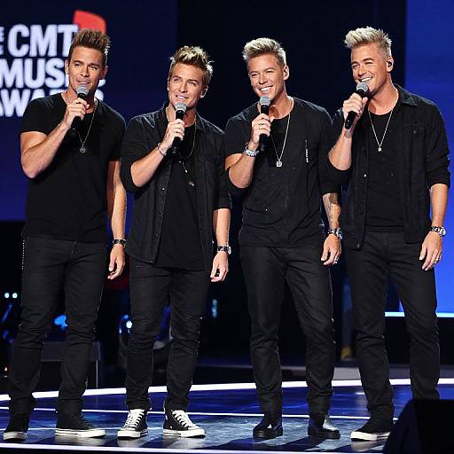 Photograph of four muscular, blonde, tattooed men in black attire, holding microphones, smiling on stage at a 