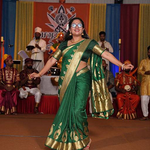 Photograph of a South Asian woman dancing on stage in a green saree with gold trim, wearing reflective sunglasses, in front of musicians and audience.