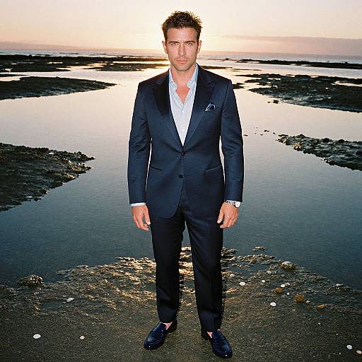 Photograph of a handsome man with short brown hair, wearing a black suit, white shirt, and black shoes, standing on a rocky shoreline at sunset