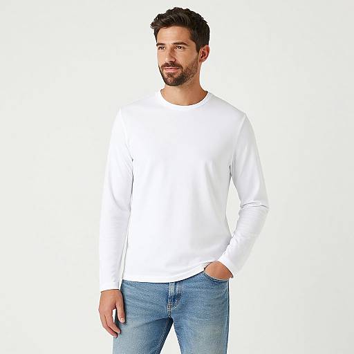 Photograph of a handsome, bearded man with short dark hair, wearing a white long-sleeve shirt and blue jeans, standing against a plain