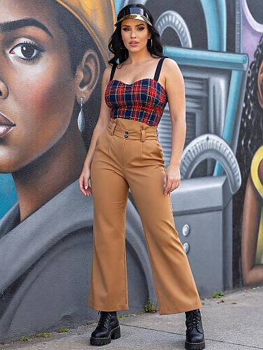 Photograph of a woman with light brown skin, wearing a black cap, plaid tank top, tan high-waisted pants, and black boots