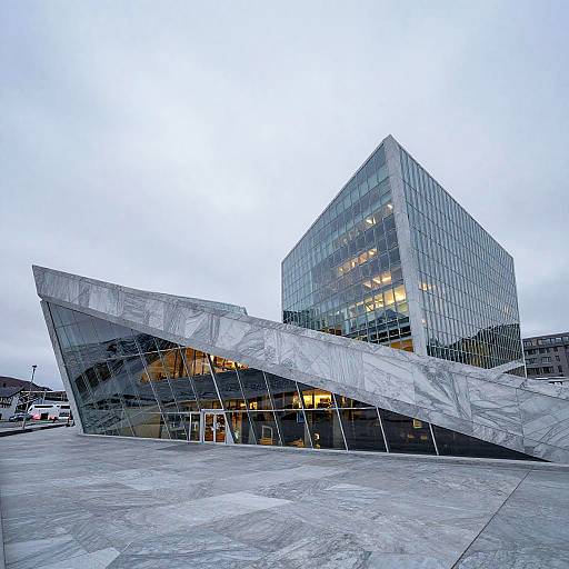 Photograph of modern, angular, glass-wrapped building with large, tilted metallic panels, illuminated windows, and concrete plaza under overcast sky.