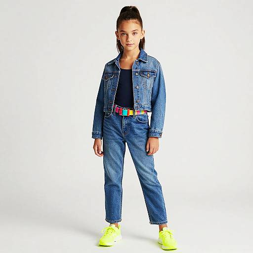 Photograph of a young woman with high ponytail, wearing a blue denim jacket, black top, colorful belt, and yellow sneakers, standing against a