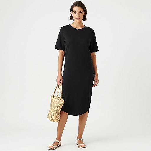 Photograph of a short-haired Asian woman in a black short-sleeve dress, holding a woven straw handbag, wearing white sandals, standing against