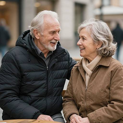 Photograph of an elderly white couple with gray hair, laughing outdoors; man in black puffer jacket, woman in brown coat.