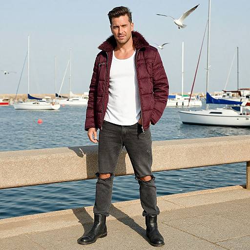 Photograph of a handsome, bearded man with short brown hair, wearing a maroon puffer jacket, white tee, ripped black jeans, and