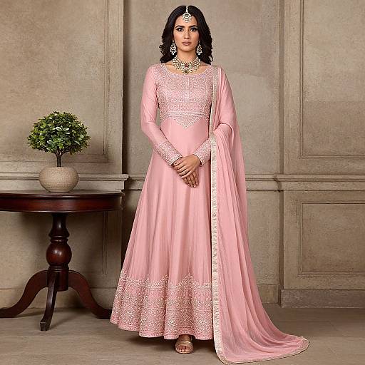 Photograph of an Indian woman in a pink silk lehenga with gold embroidery, standing against a beige paneled wall, wearing a maang tikka