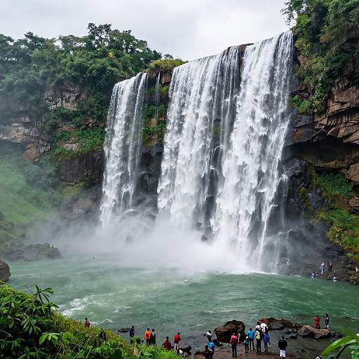 Photograph of a powerful waterfall cascading over a lush, green cliff into a misty, turquoise pool, with several people in colorful clothing standing at