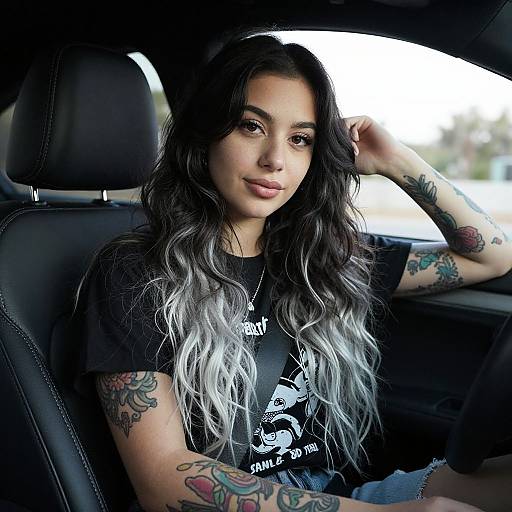 Photograph of a tattooed woman with long, ombre black-to-gray hair, wearing a black graphic tee, seated in a car, smiling