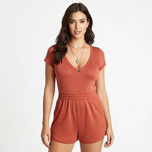 Photograph of a woman with long black hair, wearing a red V-neck romper, layered necklaces, standing against a plain white background.